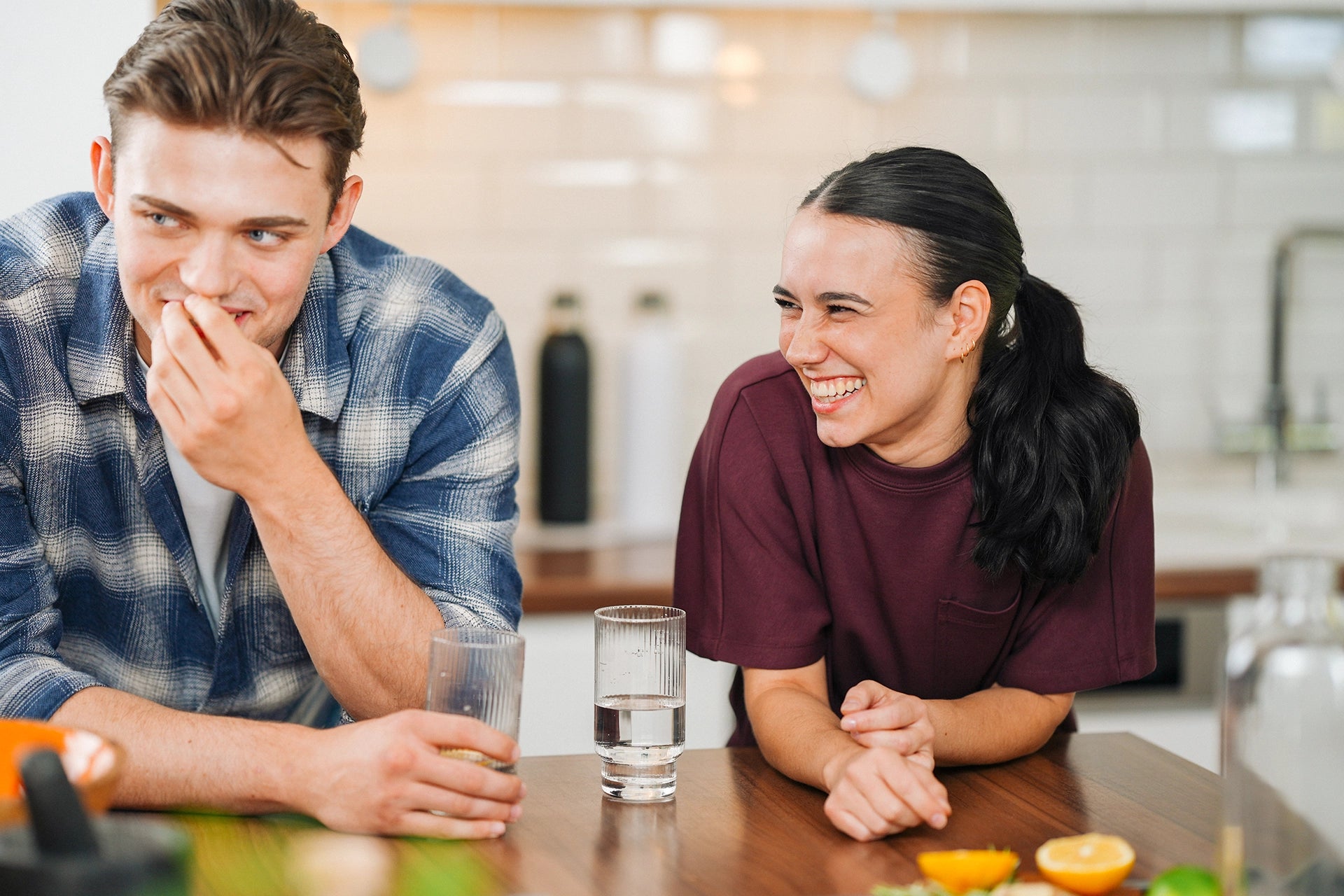Two young people laughing together at a kitchen table with glasses of water.