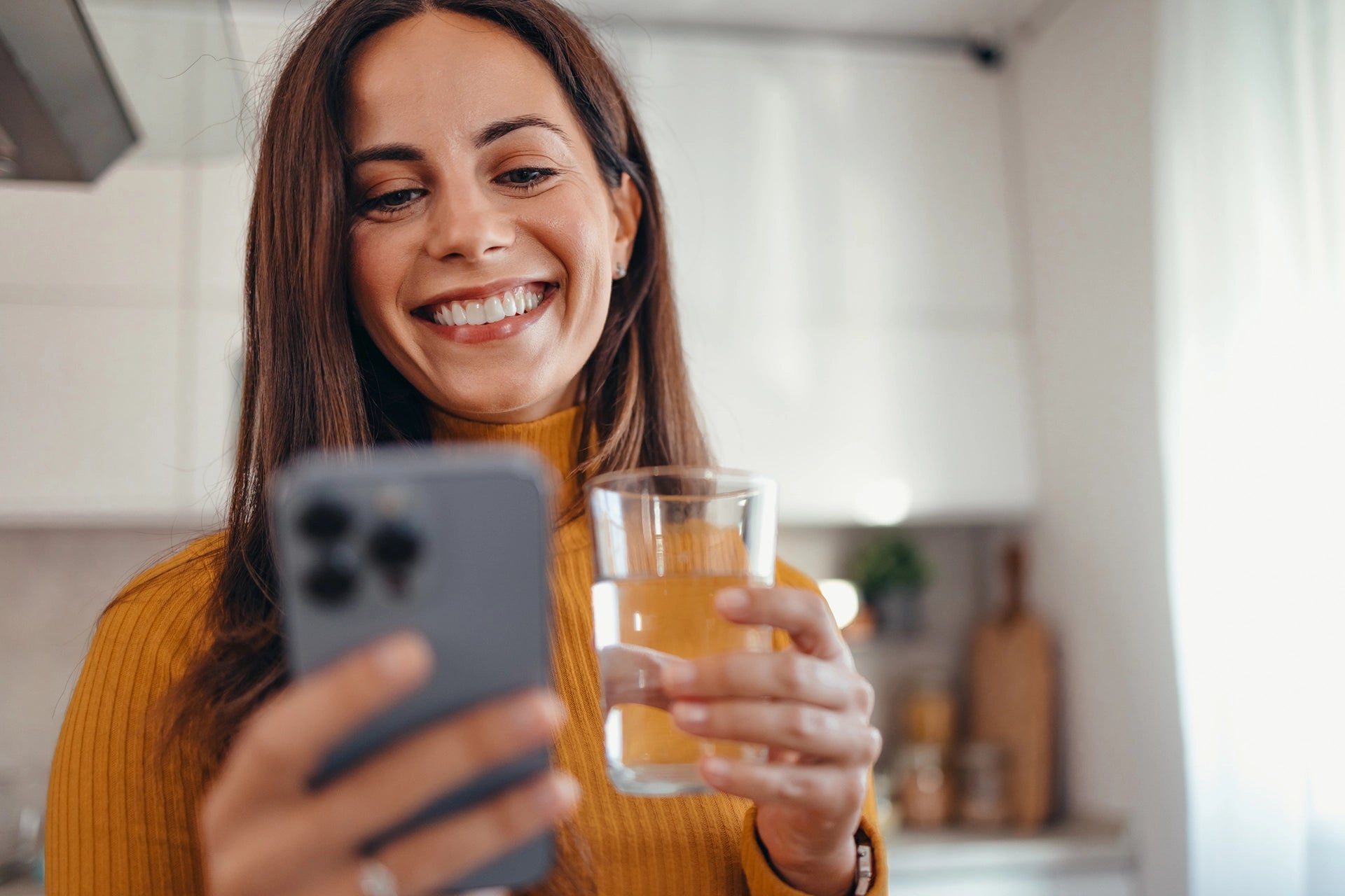Woman in a kitchen holding a glass of water and a smartphone.