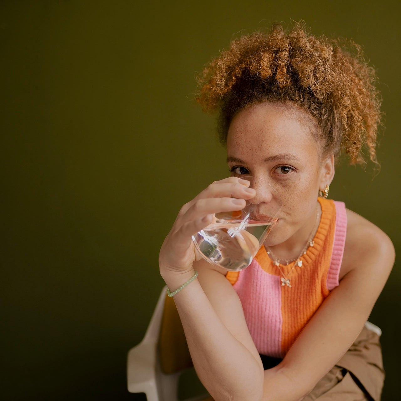 Woman in pink shirt drinks water in front of a green wall.