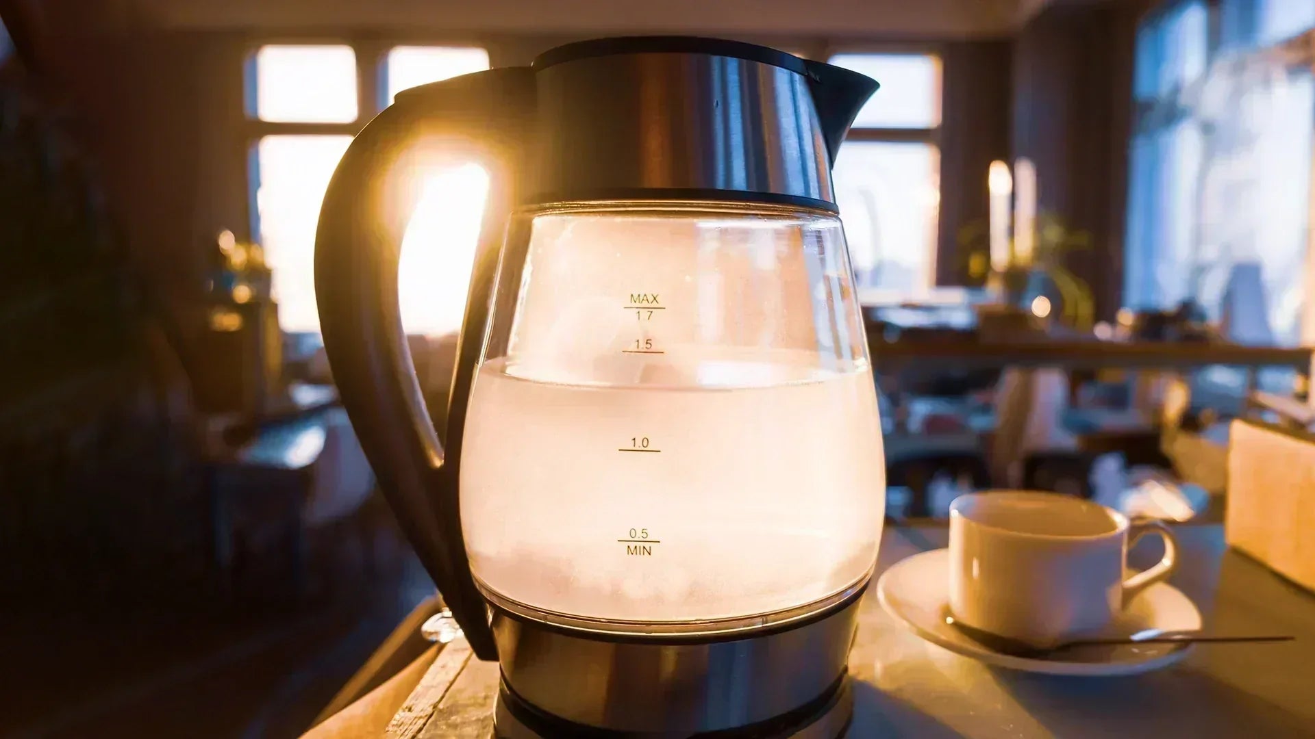 A glass electric kettle, filled with cloudy tap water, shown on a table in a warm indoor setting. A white cup sits on a saucer to the right.
