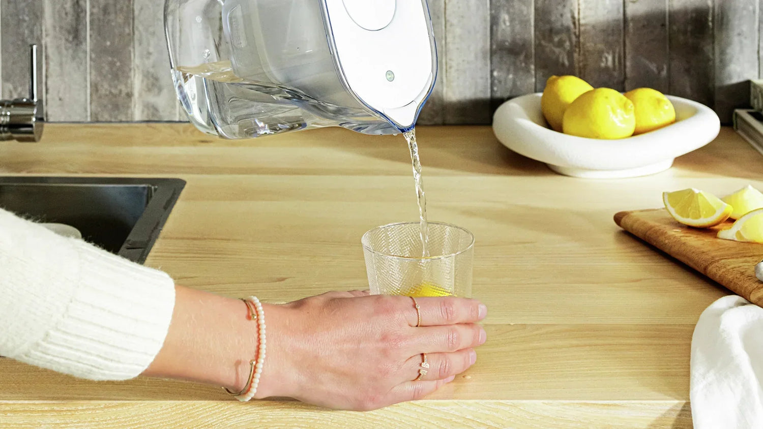 A woman fills in a glass of water with a water jug in a kitchen. In the background stands a bowl with bright yellow lemons.