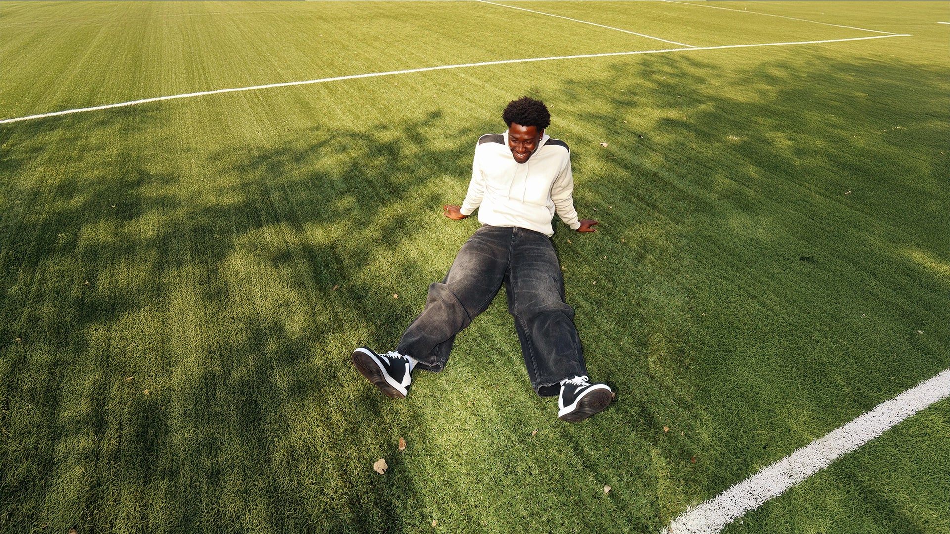 A young man sitting on a grass sports field, wearing a light-coloured hoodie, dark jeans and black trainers, with white field markings visible in the background.