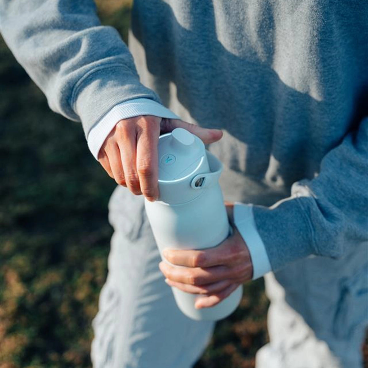 A close-up of a person outdoors holding and opening a white bottle with a flip-top lid, wearing a gray sweatshirt and light gray pants.