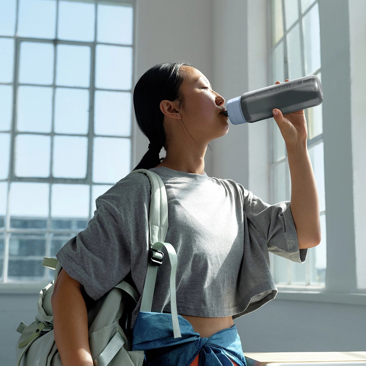 Una donna che beve da una caraffa BRITA filtro Modello "Bottle" in una stanza luminosa con grandi finestre.