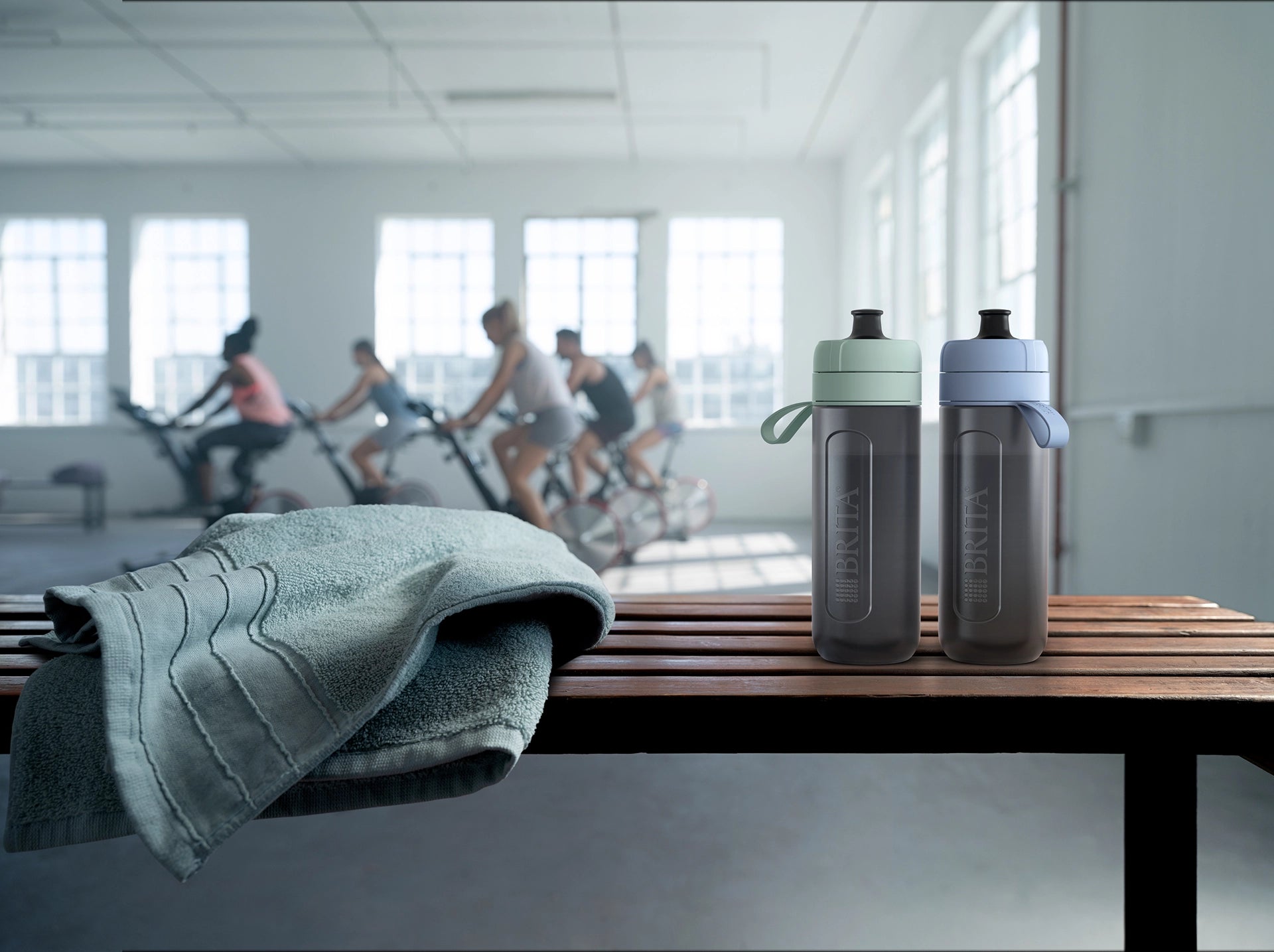 Two BRITA water filter bottles Model Active and a towel on a bench with people exercising in the background