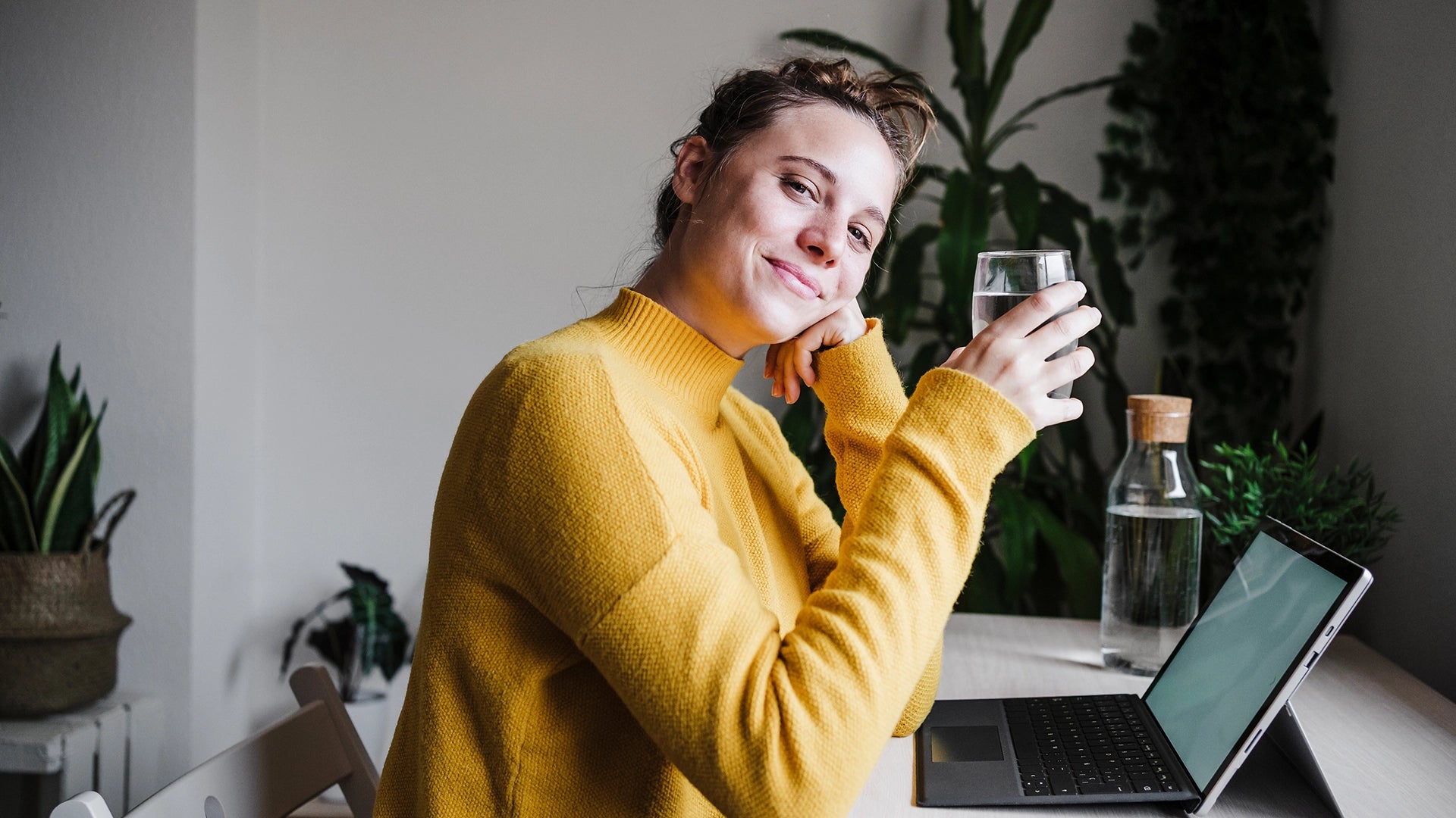 A young woman with a yellow shirt is drinking a glass of water at her desk.