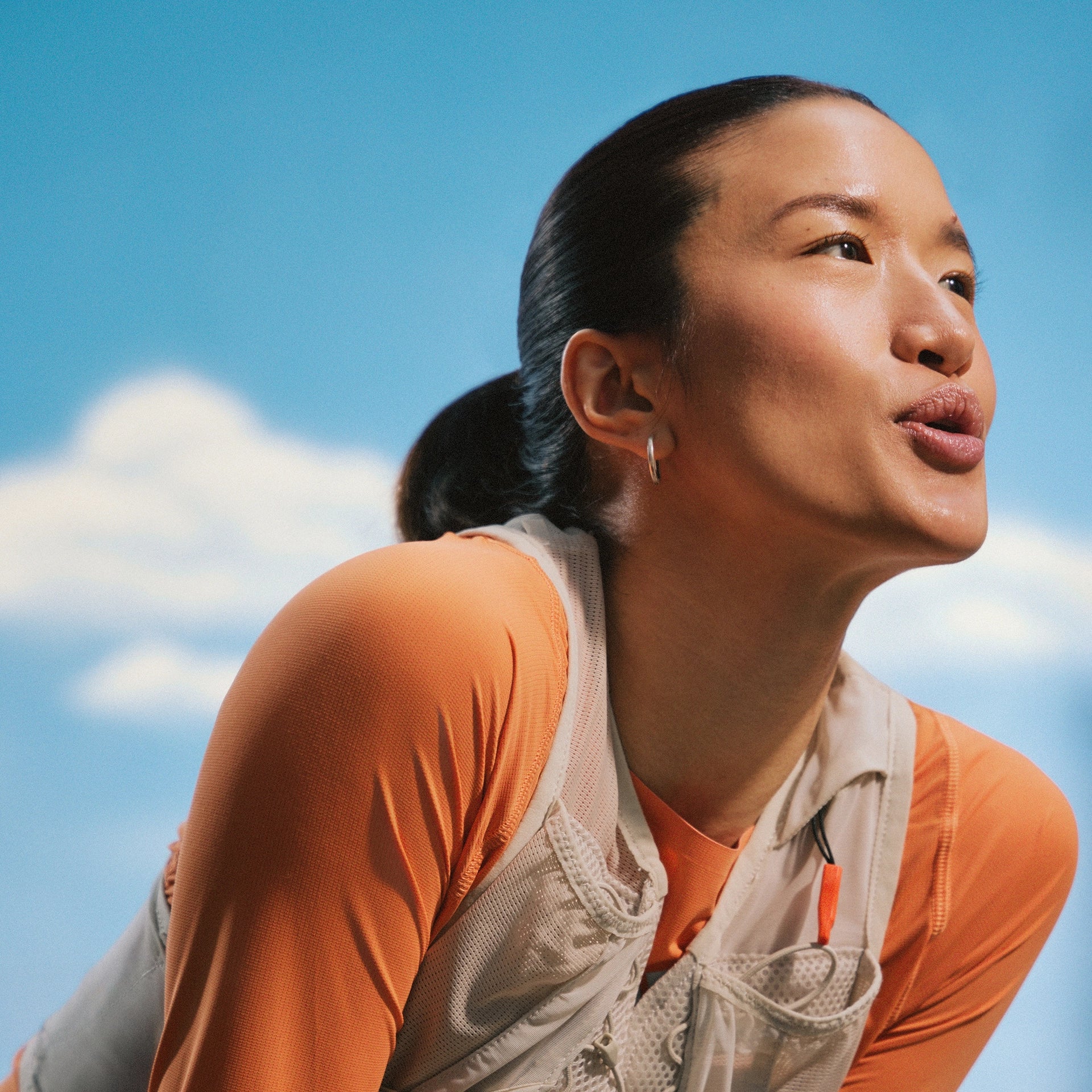 Woman wearing an orange shirt and beige apron against a blue sky with clouds.