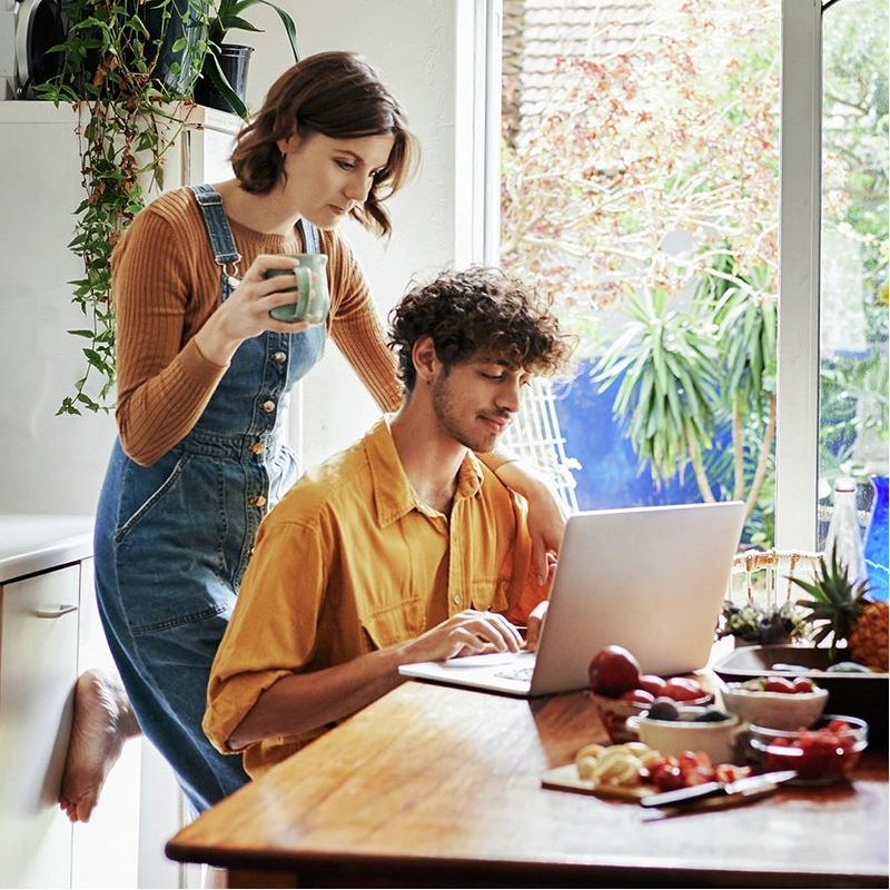 Woman holding a mug while standing beside a man working on a laptop at a wooden table in a bright kitchen with plants, flowers, and fresh fruit.