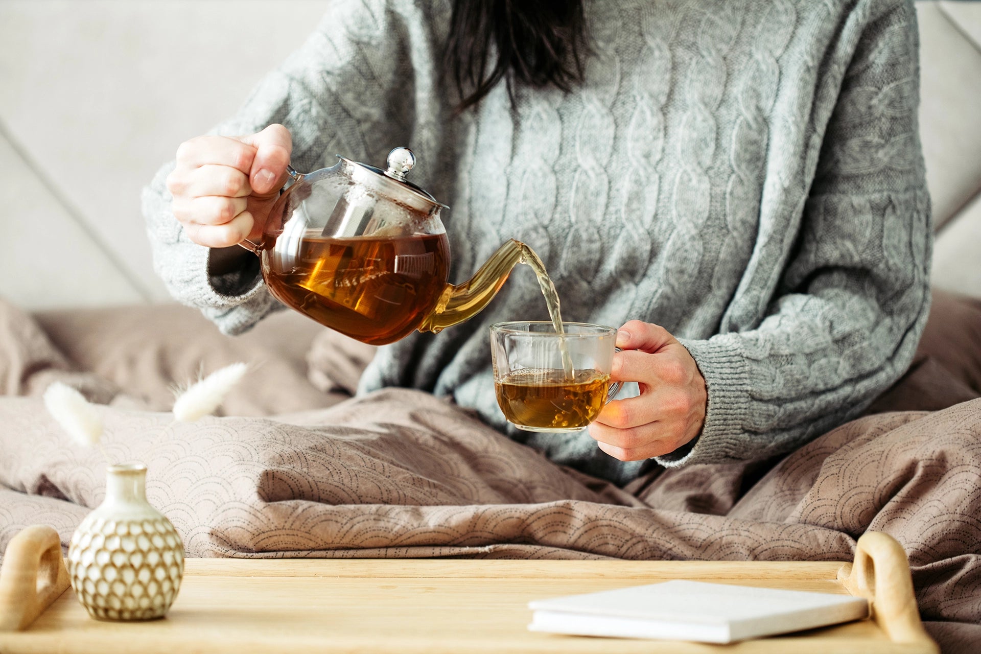 Person pouring tea from a glass teapot into a cup on a wooden table.