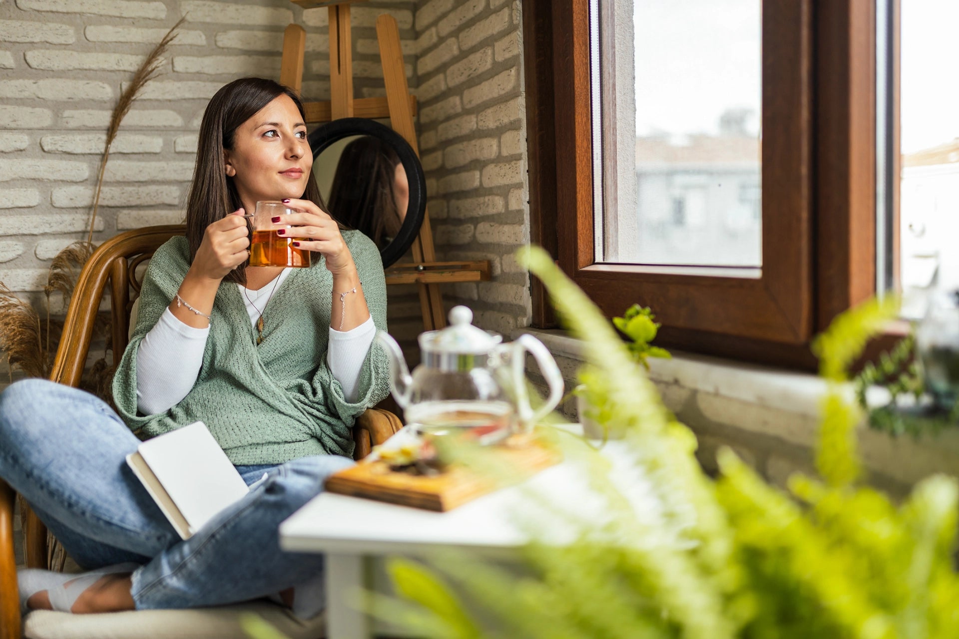 Woman sitting on a chair holding a mug, with a table in front of her containing a teapot and books, in a cozy room.
