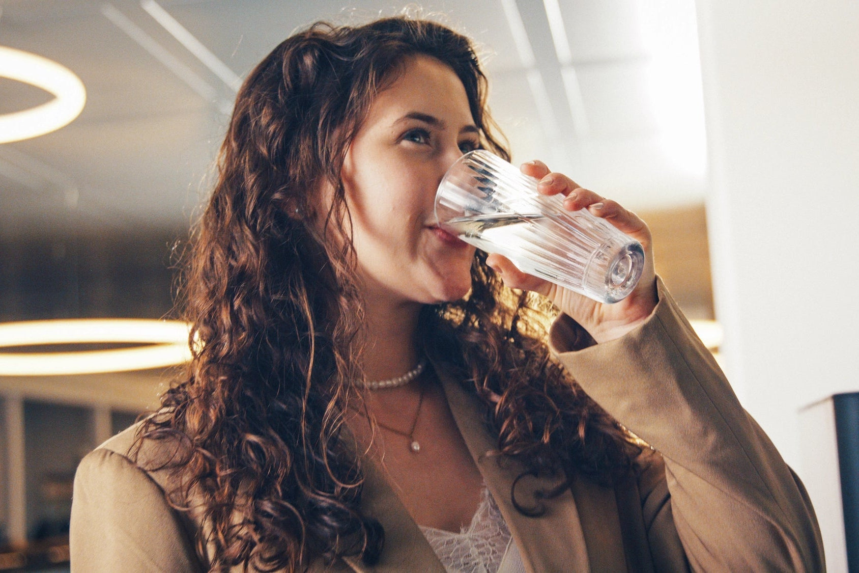 A woman with long curly brown hair drinking water from a glass in an indoor setting.