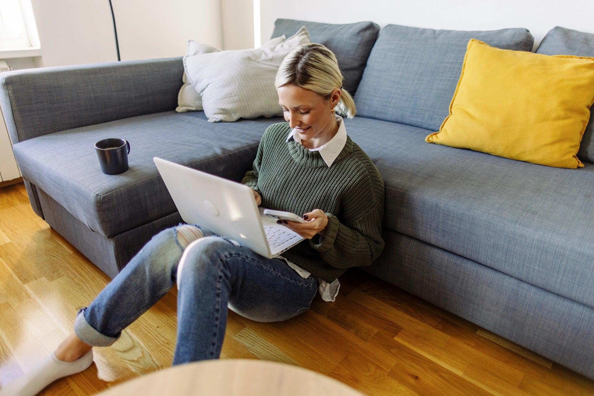 Woman sitting on the floor using a laptop, with a cup on a sofa nearby.