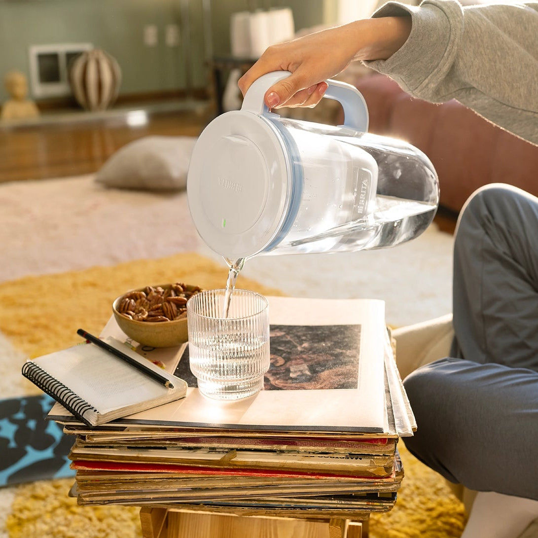Person pouring filtered water from a blue BRITA Glass Jug into a glass on a table with books and snacks.