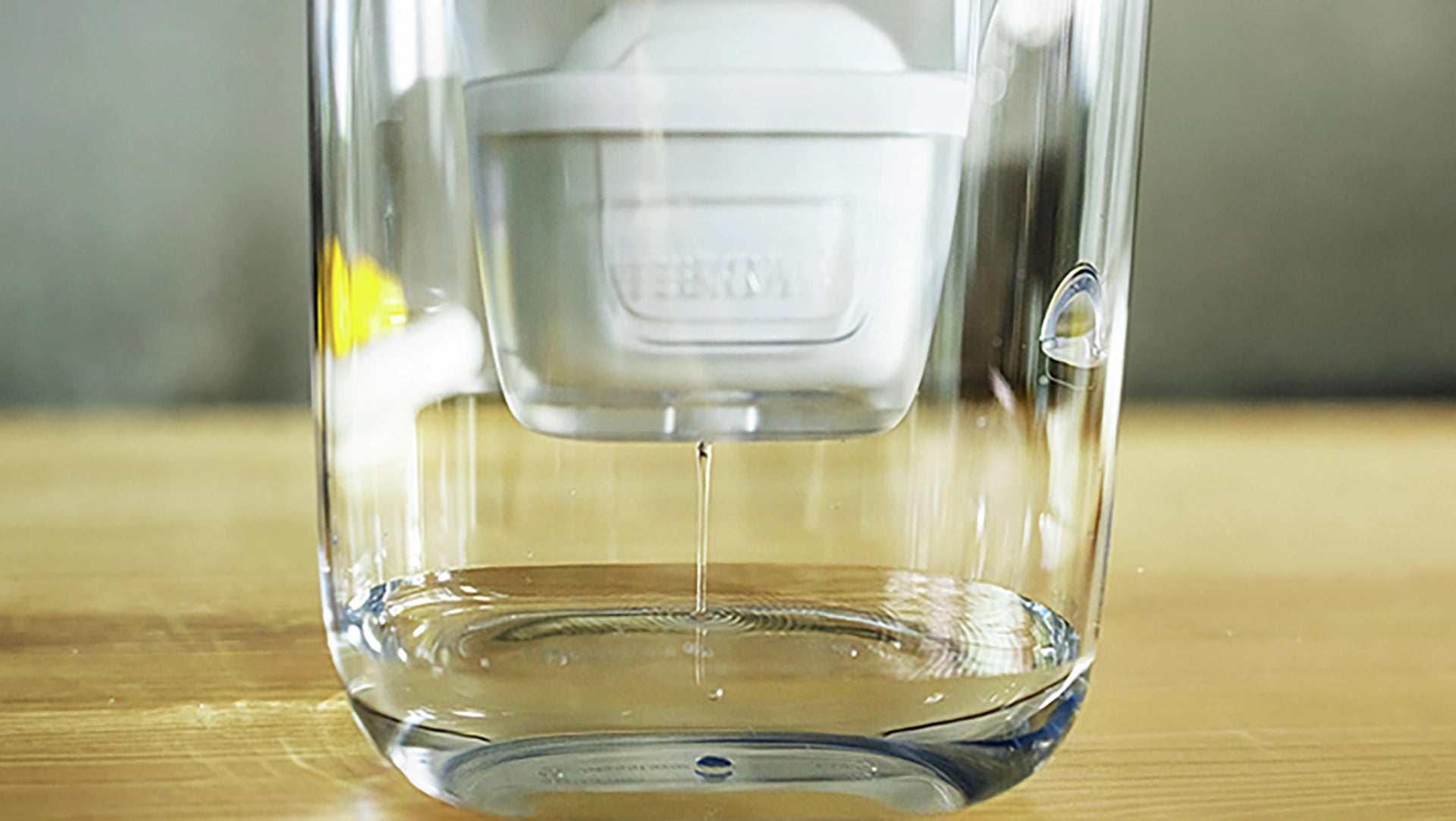 Close-up of a BRITA MAXTRA water filter cartridge inserted in a jug, with filtered water dripping into a clear glass container
