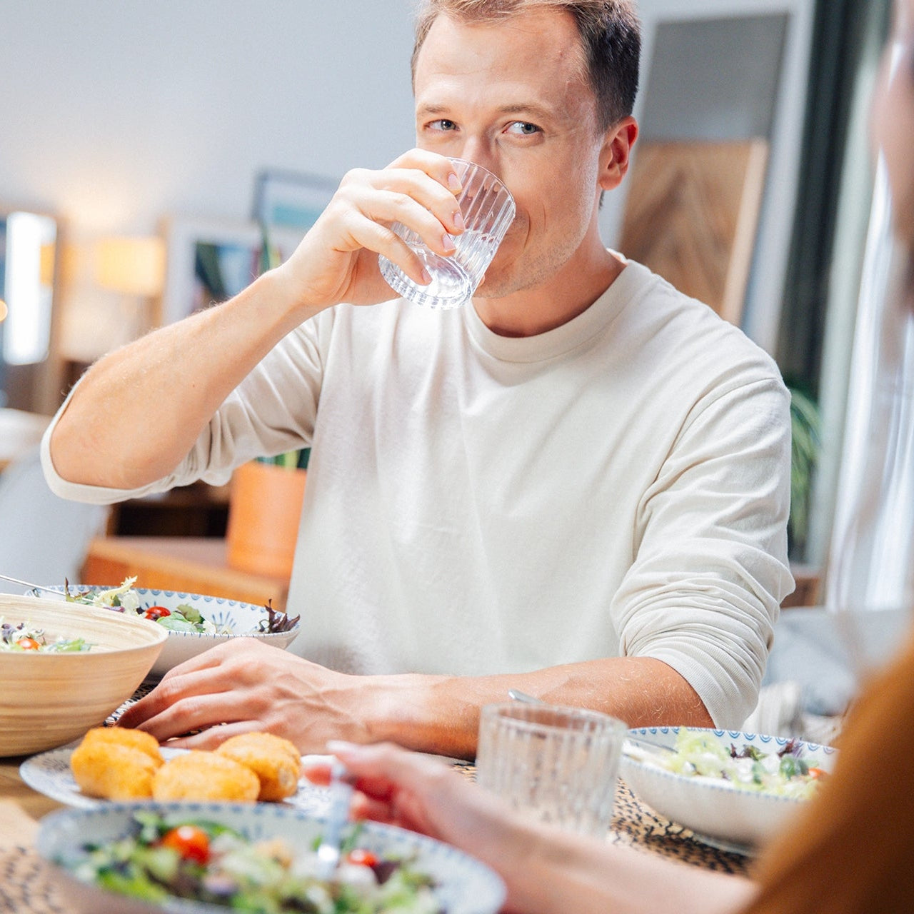 Man drinking water at a dining table with food and another person partially visible