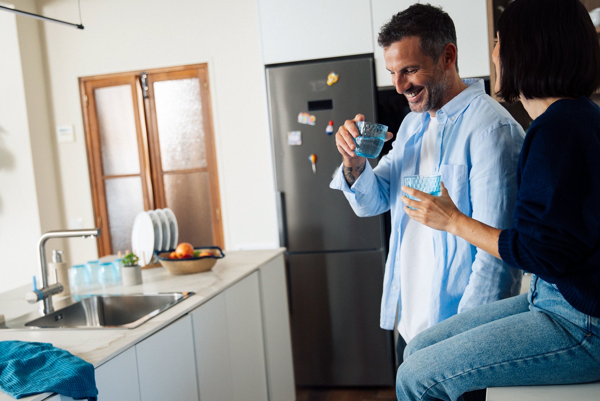 Man and Woman holding glasses of water in a modern kitchen beside a 3-way-tap and sink.