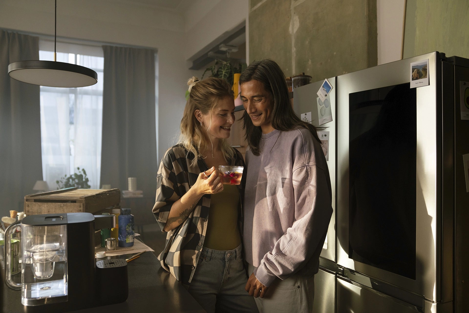 Two people standing in a kitchen next to a BRITA YOURCE mini water dispenser, with one person holding a glass cup of tea.