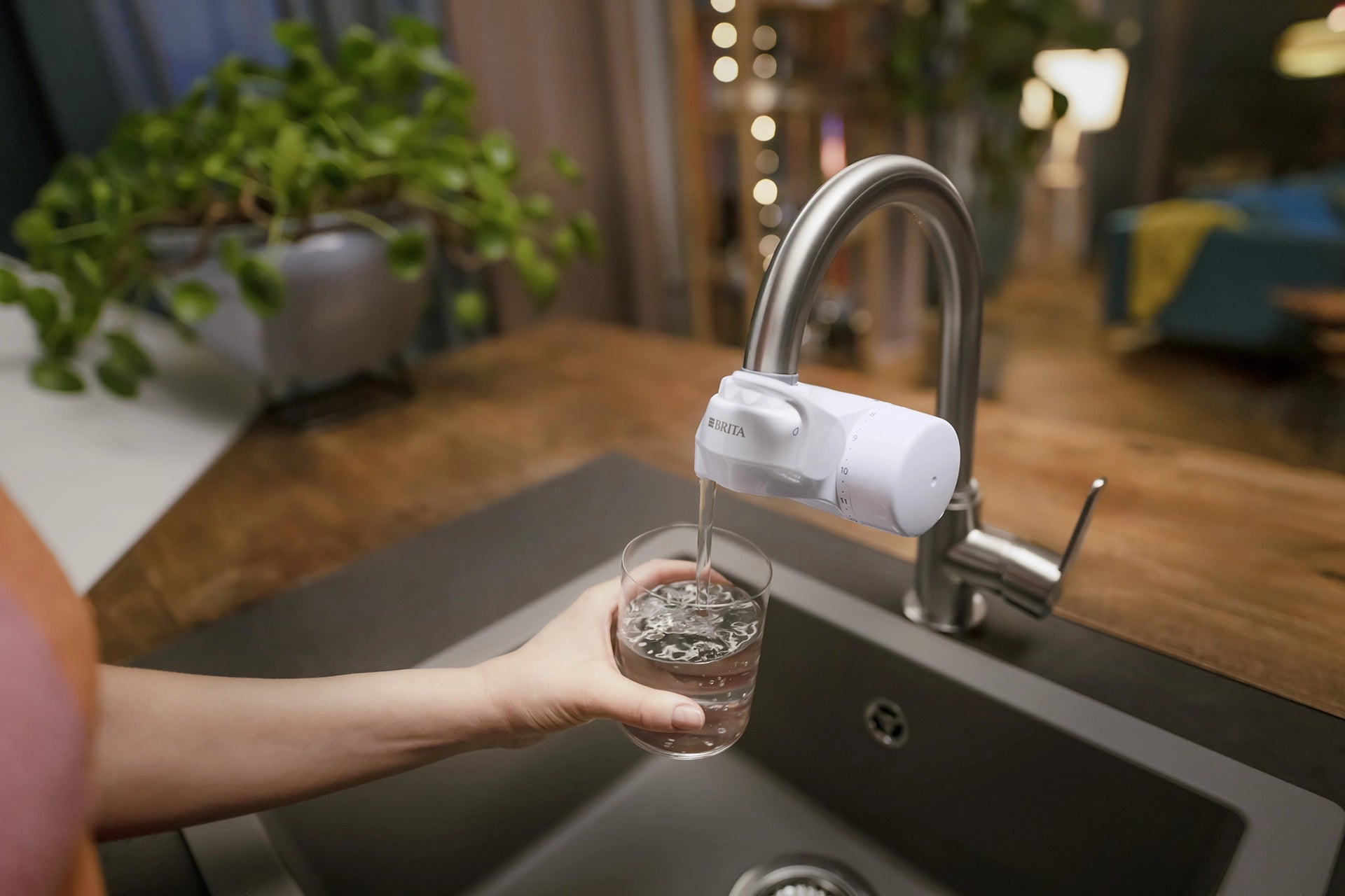Person filling a glass with filtered water from a BRITA ON TAP water filter at a kitchen sink.